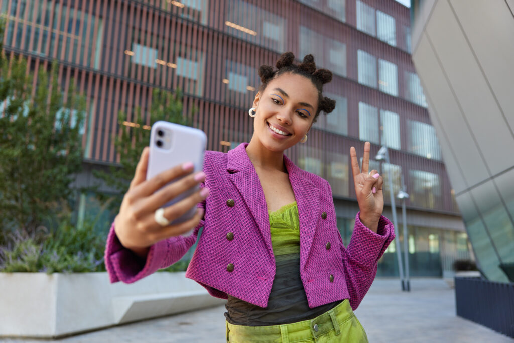 Attractive Girl In Fashionable Outfit Creats Influence Content Shows Peace Sign At Smartphone Front Camera Poses For Selfie Smiles Gladfully Poses At Urban Place Spends Vacation At Big City. - PAVON | Contabilidade em São Paulo