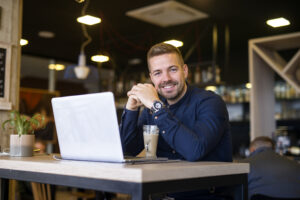 Portrait Of Smiling Man Sitting In A Cafe Bar With His Laptop Computer. - PAVON | Contabilidade em São Paulo