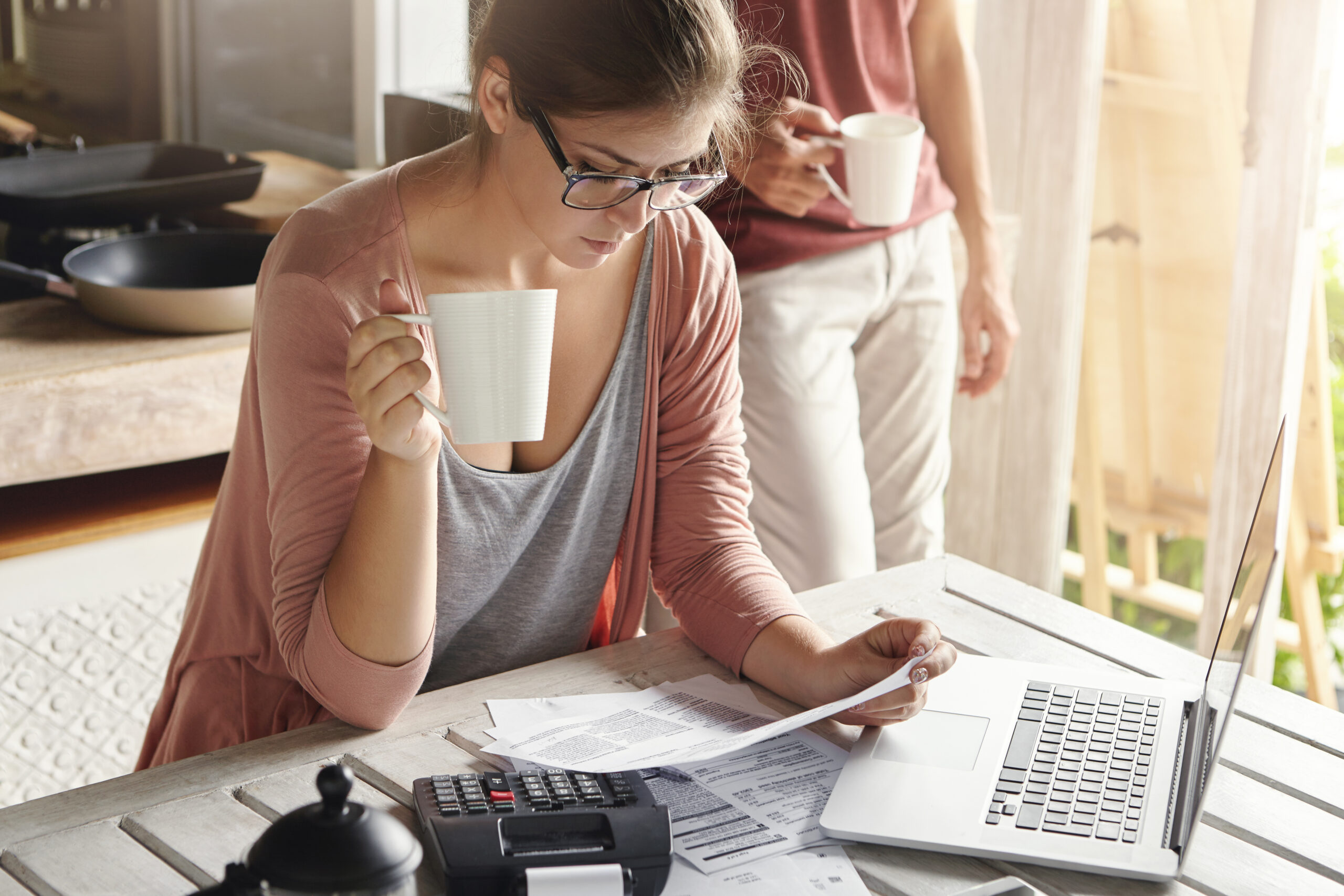 Young Female Drinking Tea And Studying Bill In Her Hands, Having Frustrated Look While Managing Family Budget And Doing Paperwork, Sitting At Kitchen Table With Papers, Calculator And Laptop Computer - PAVON | Contabilidade em São Paulo