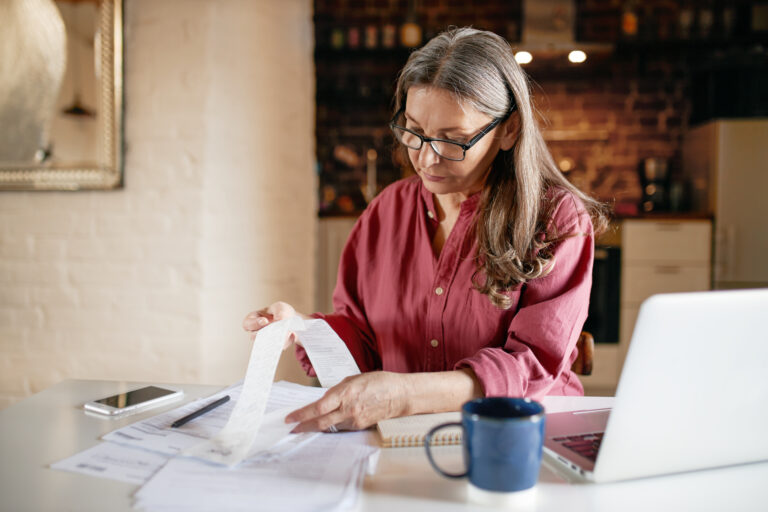 Serious Focused Middle Aged Female Accountant In Eyewear Working From Home Providing Outsource Bookkeeping Service, Sitting At Table With Portable Computer, Calculating Finances, Doing Paperwork - PAVON | Contabilidade em São Paulo