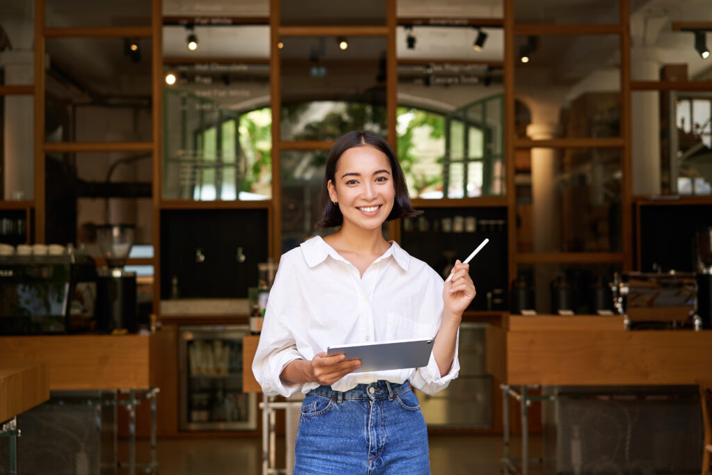 Portrait Of Asian Woman, Manager Standing With Tablet In Front Of Cafe Entrance, Welcomes Guests - PAVON | Contabilidade em São Paulo