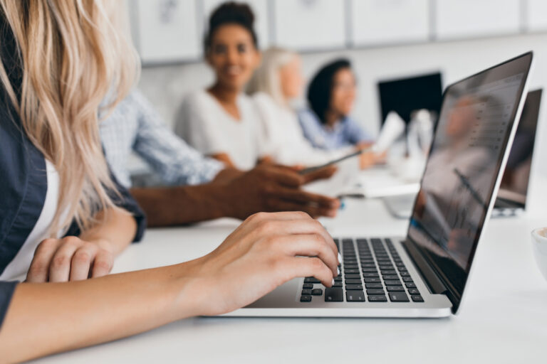 Blonde Woman With Elegant Hairstyle Typing Text On Keyboard In Office. Indoor Portrait Of International Employees With Secretary Using Laptop On Foreground.. - PAVON | Contabilidade em São Paulo