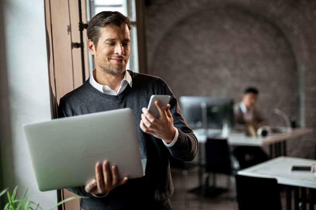 Happy Entrepreneur Texting On Smart Phone While Holding Laptop In The Office. - PAVON | Contabilidade em São Paulo