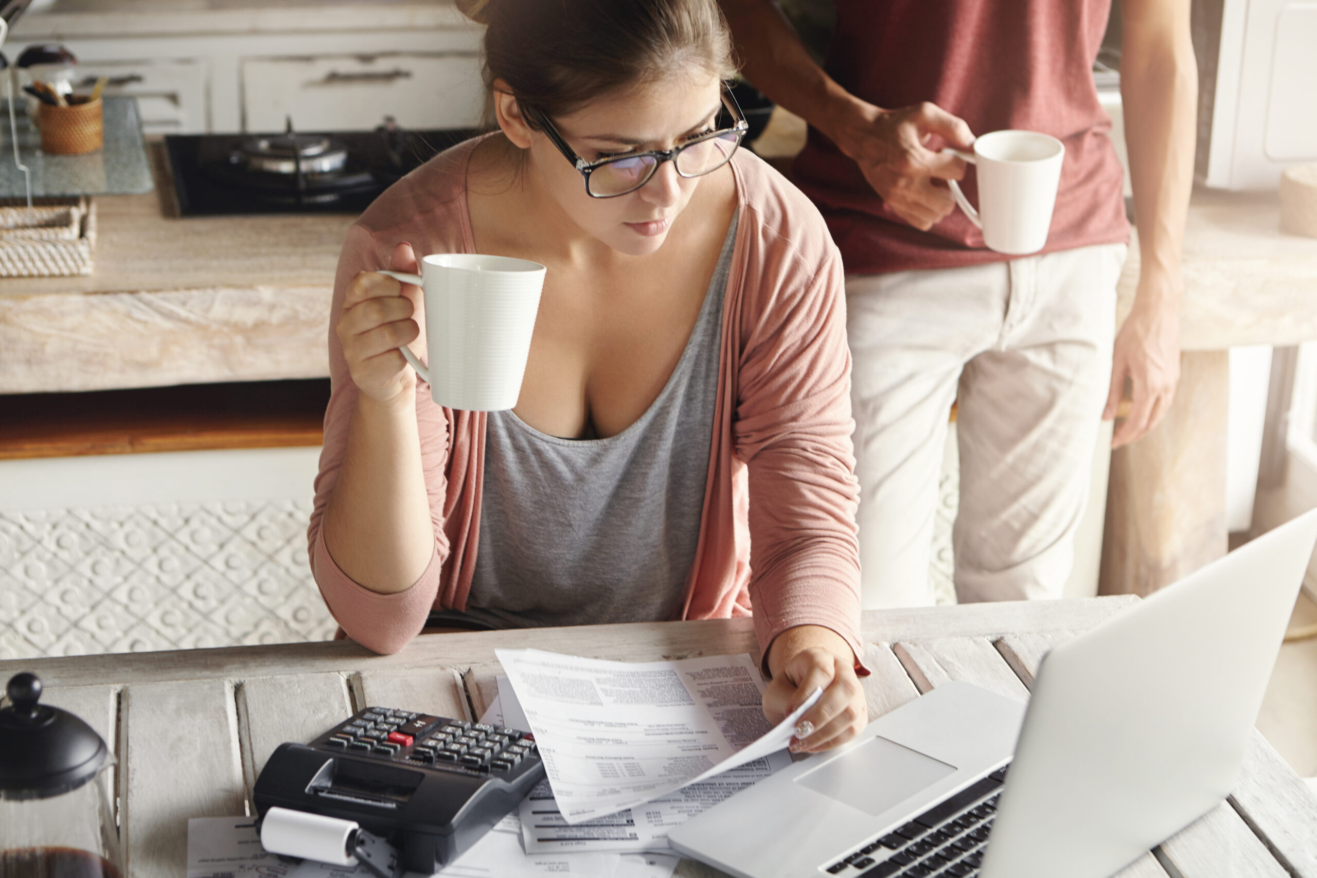 Young Couple Calculating Family Expenses At Home. Woman In Spectacles Paying Utility Bills Online, Having Coffee Or Tea, Sitting In Kitchen With Documents And Calculator, Looking At Laptop Screen - PAVON | Contabilidade em São Paulo
