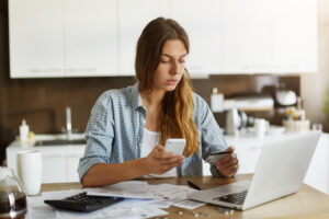 Indoor Portrait Of Serious Attractive Woman Holding Debit Card And Cell Phone, Entering Data Via Online Payment Form While Ordering Food Delivery Or Shopping, Sitting In Kitchen, Using Laptop Pc - PAVON | Contabilidade em São Paulo