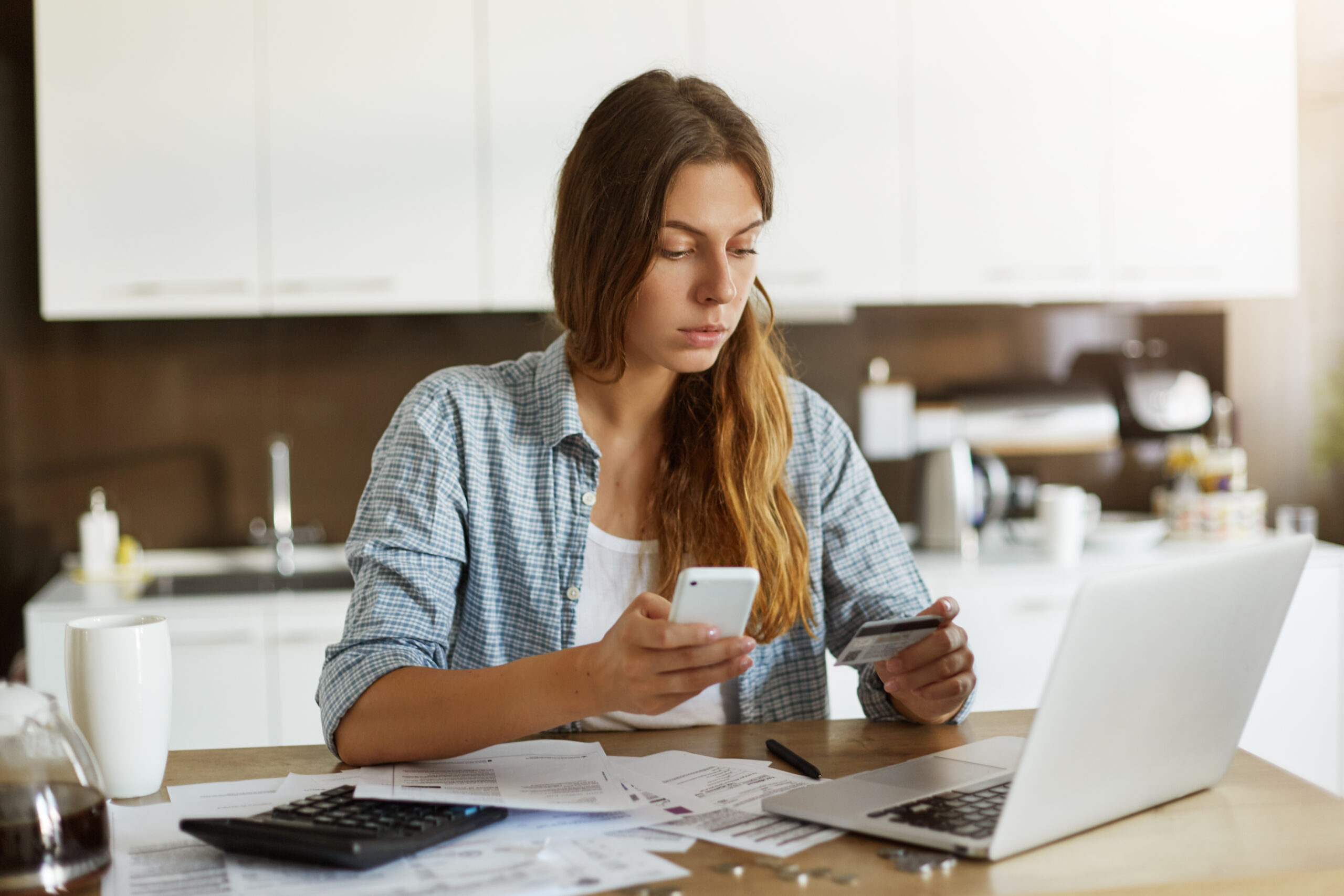 Indoor Portrait Of Serious Attractive Woman Holding Debit Card And Cell Phone, Entering Data Via Online Payment Form While Ordering Food Delivery Or Shopping, Sitting In Kitchen, Using Laptop Pc - PAVON | Contabilidade em São Paulo